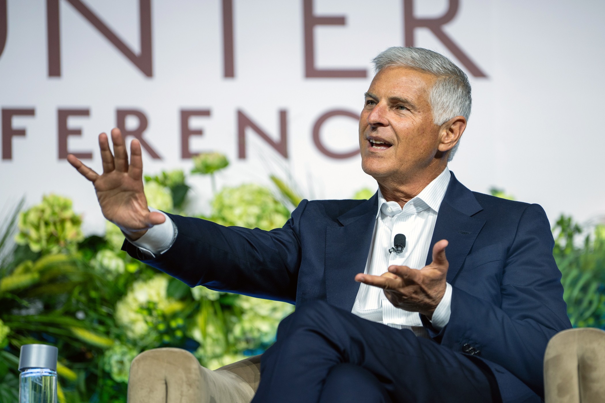 A man in dark blue suit gestures while speaking at a conference with floral backdrop and "CONFERENCE" text. A man in dark blue suit gestures while speaking at a conference with floral backdrop and "CONFERENCE" text.