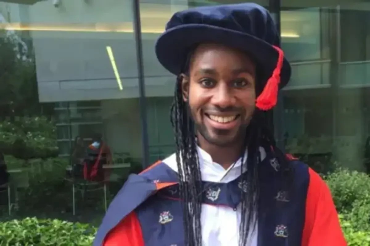 A person in academic regalia poses outdoors near a modern glass building. A person in academic regalia poses outdoors near a modern glass building.