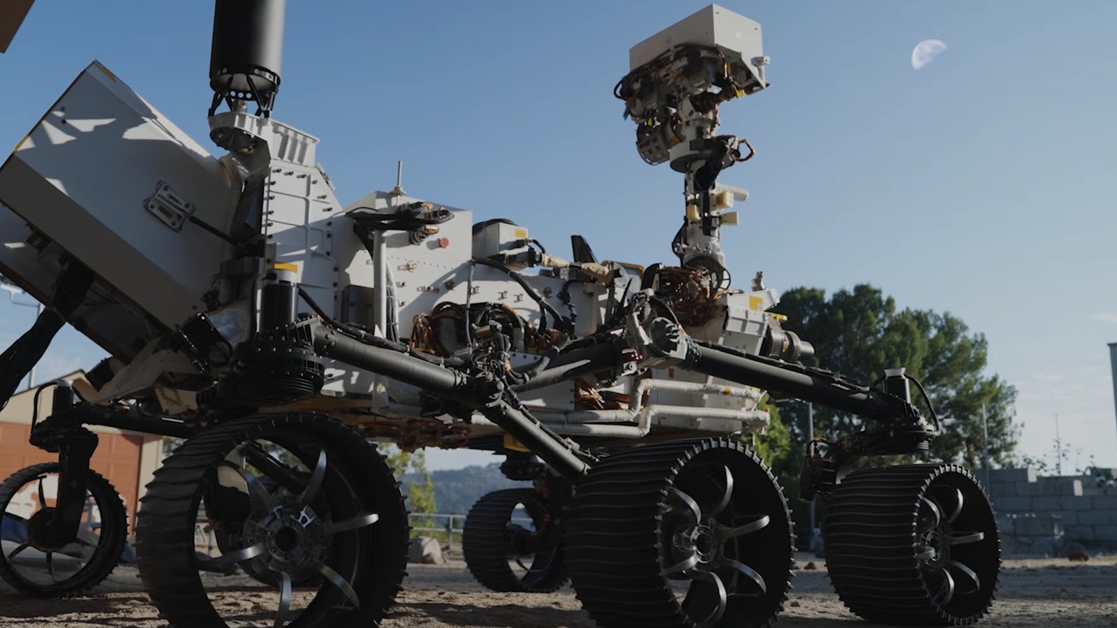 A Mars rover sits stationary on dusty ground beneath a clear blue sky with trees visible.