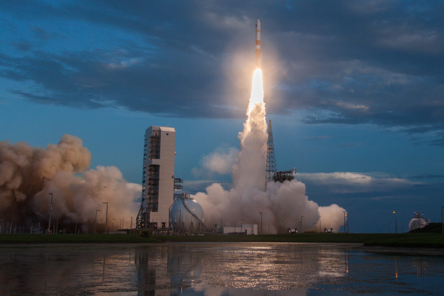 Rocket launches vertically from a pad, emitting bright flames and thick smoke under a twilight sky.