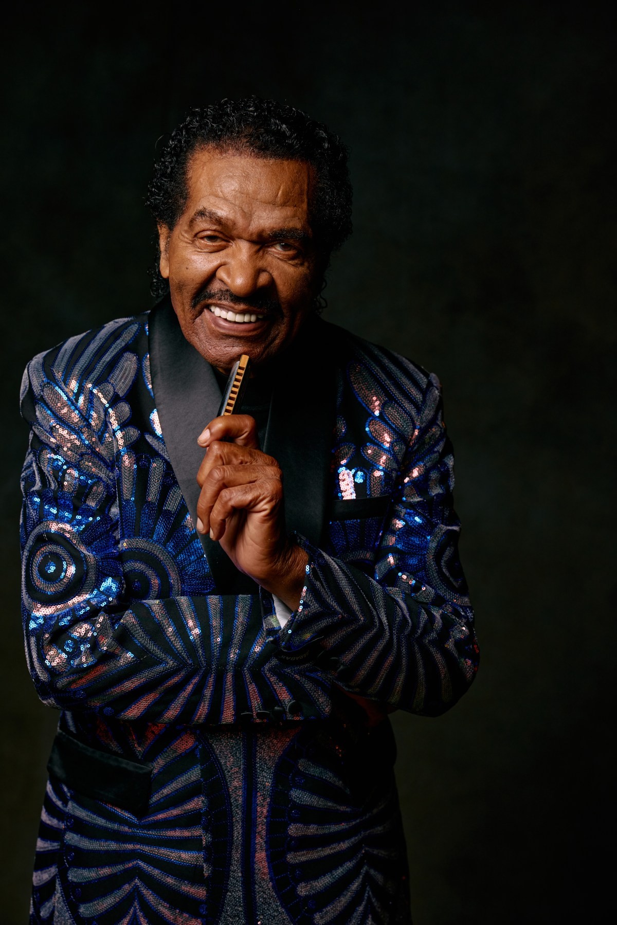 A man in a sequined blue suit holds a harmonica against a dark backdrop. A man in a sequined blue suit holds a harmonica against a dark backdrop.