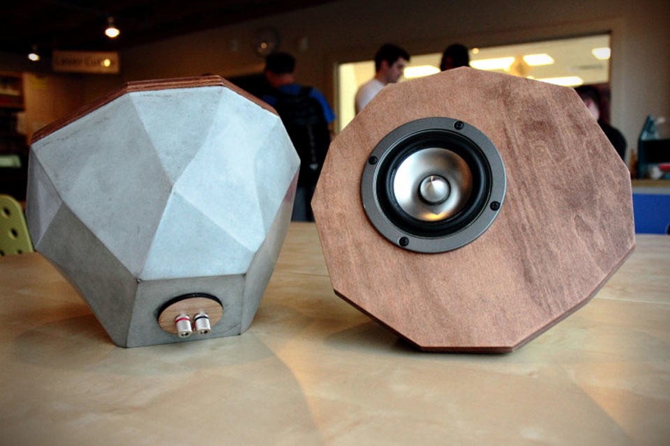 Two speakers—one concrete with inputs, one wooden with cone—rest on an indoor table.