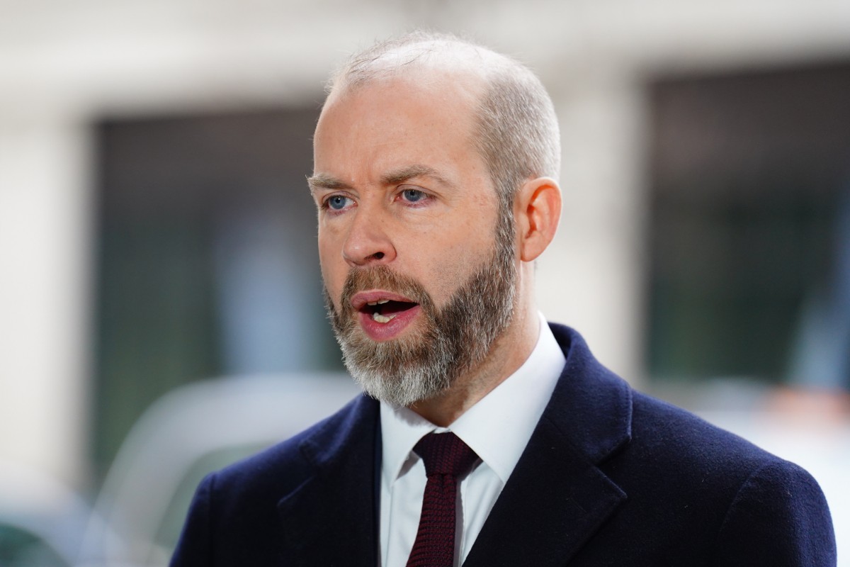 A bearded man in a dark suit and maroon tie speaks outdoors near blurred buildings.