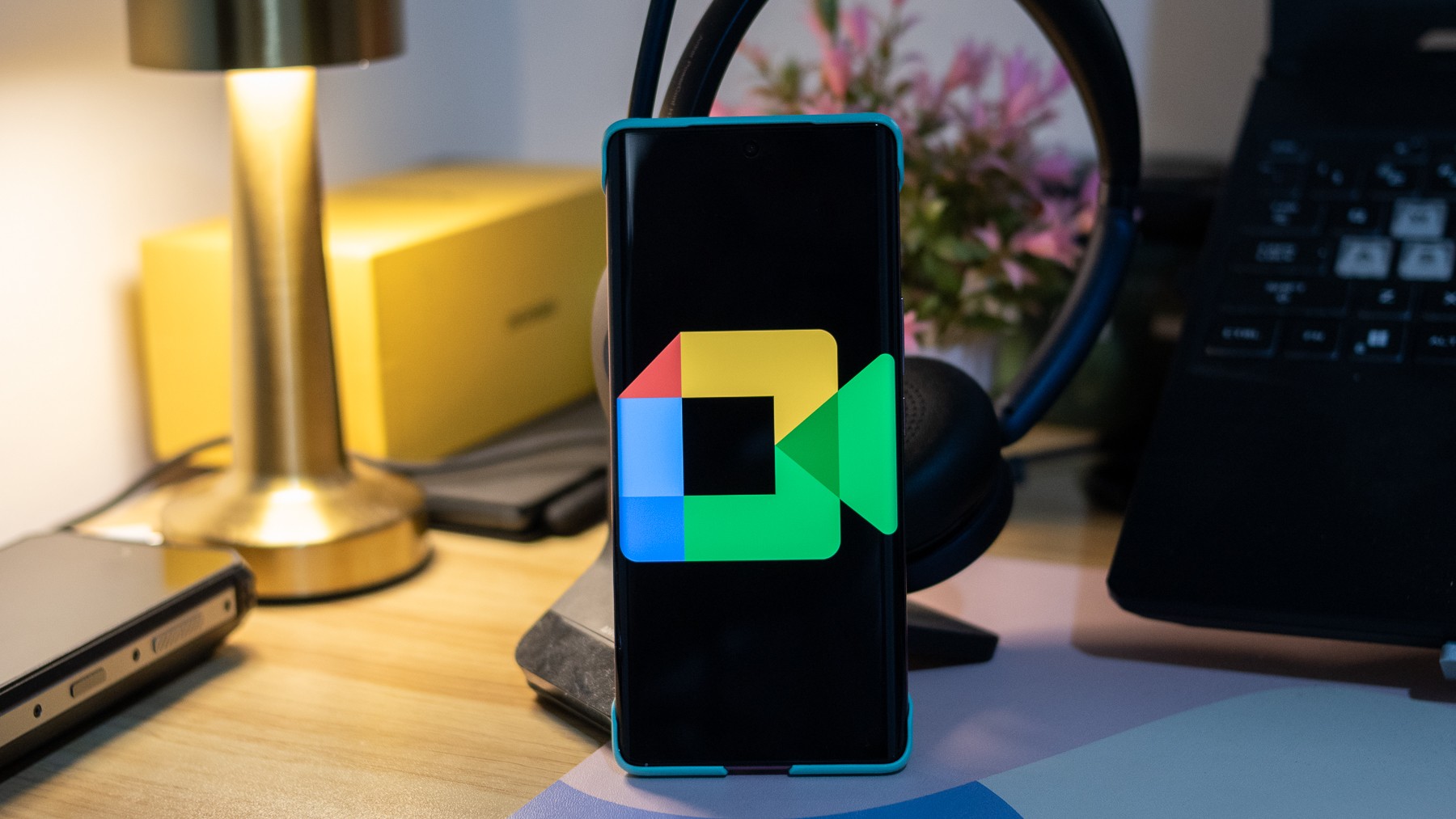 A smartphone displays the Google Meet logo on a desk with a lamp, headphones, and laptop. A smartphone displays the Google Meet logo on a desk with a lamp, headphones, and laptop.