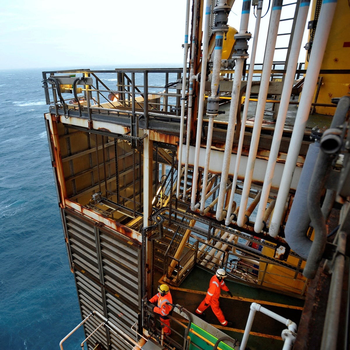 Two workers in orange suits walk on an offshore oil rig platform above the ocean. Two workers in orange suits walk on an offshore oil rig platform above the ocean.