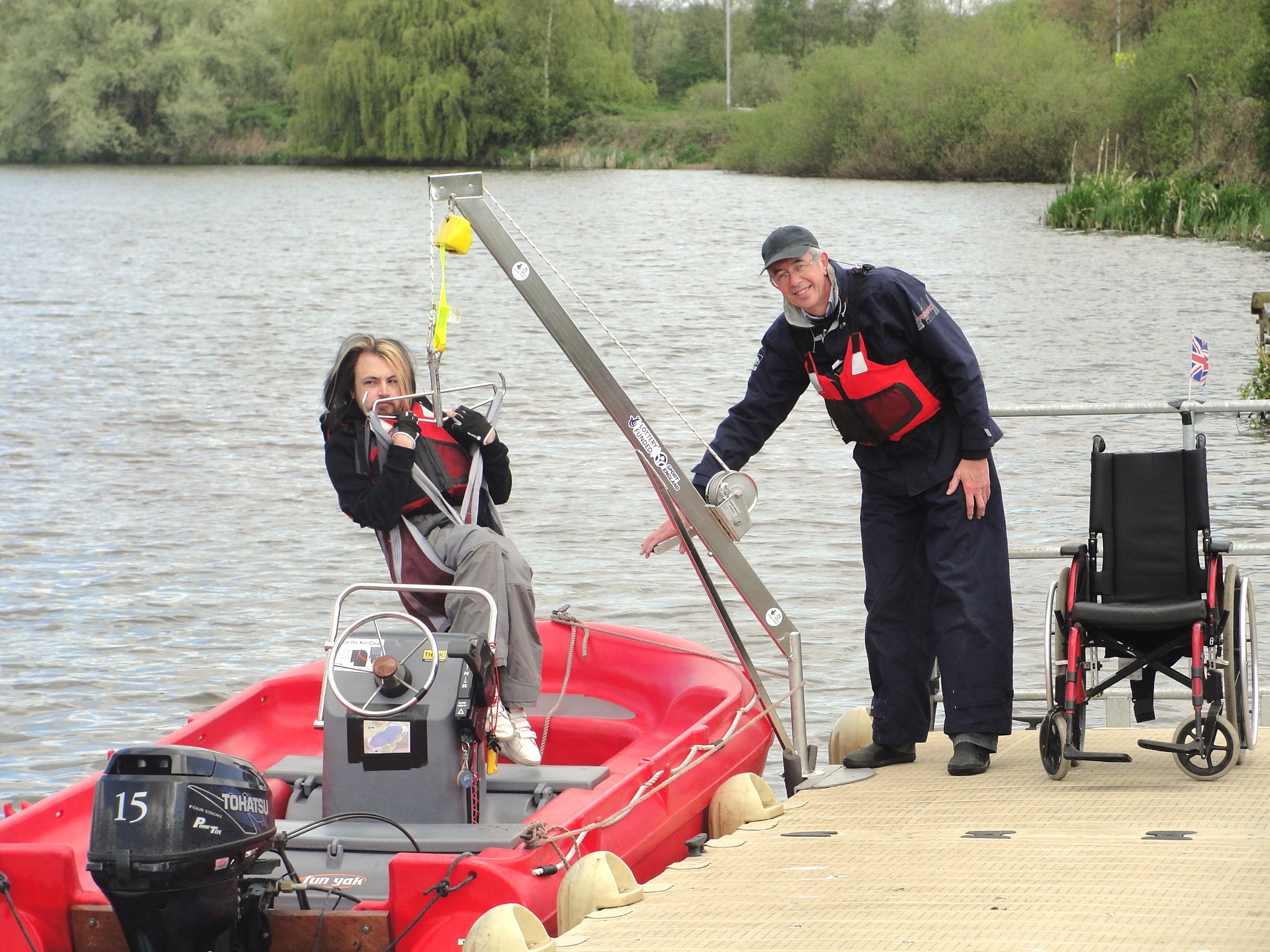 A person in a red boat is assisted by another at a riverside dock near a wheelchair. A person in a red boat is assisted by another at a riverside dock near a wheelchair.