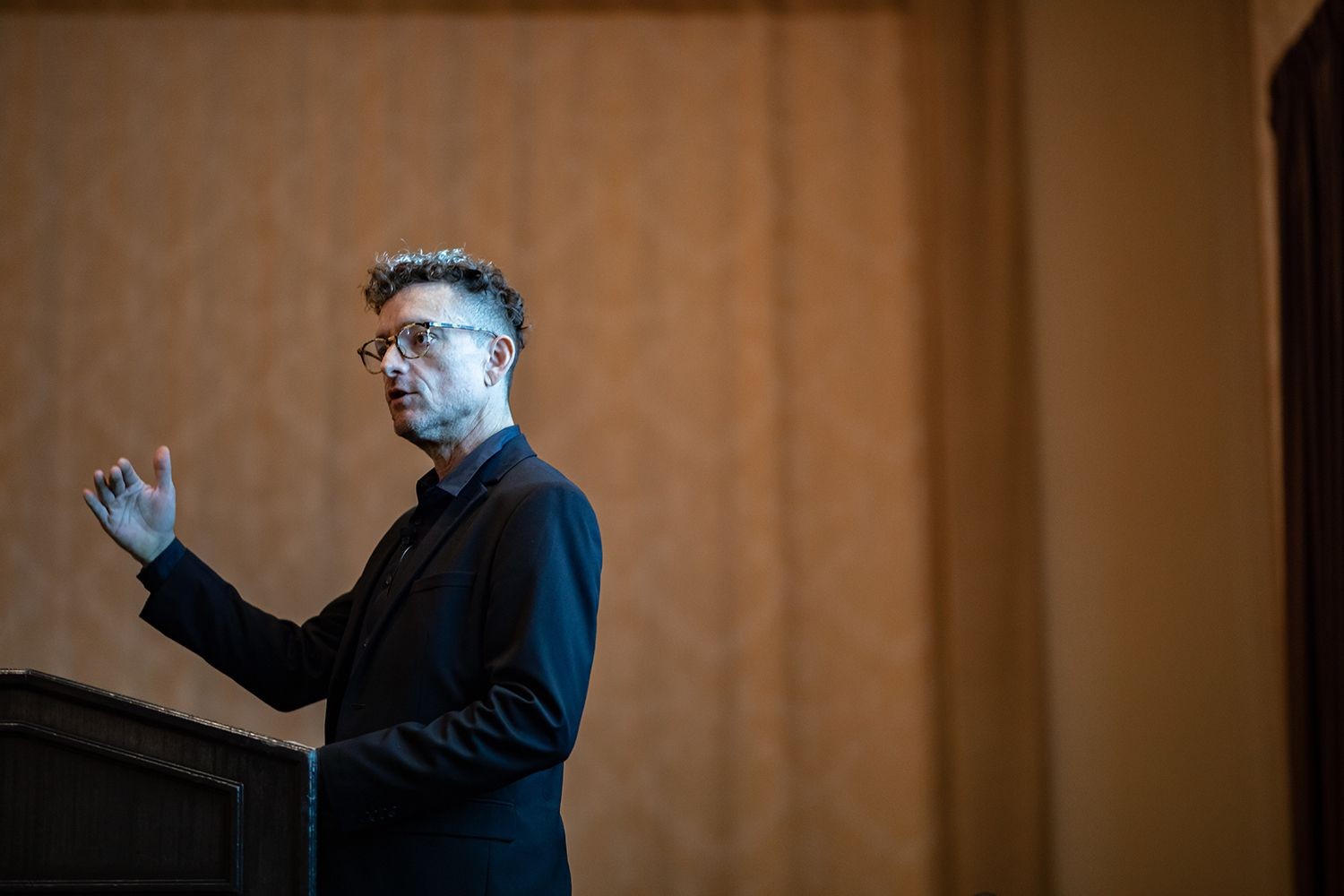 A man in a dark suit speaks at a podium against a wooden wall.