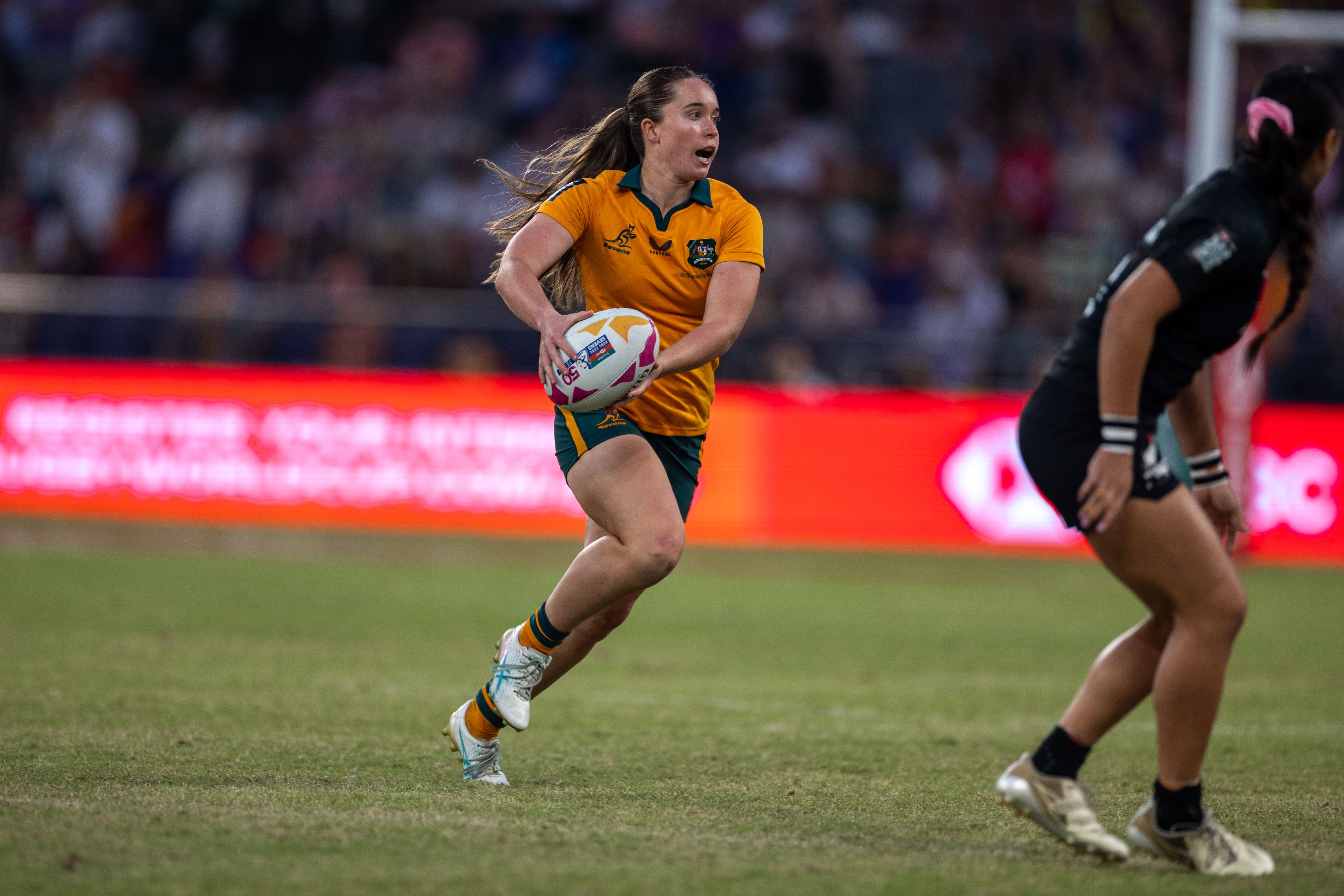 A female rugby player in an orange jersey runs with a ball on a grass field during a match. A female rugby player in an orange jersey runs with a ball on a grass field during a match.