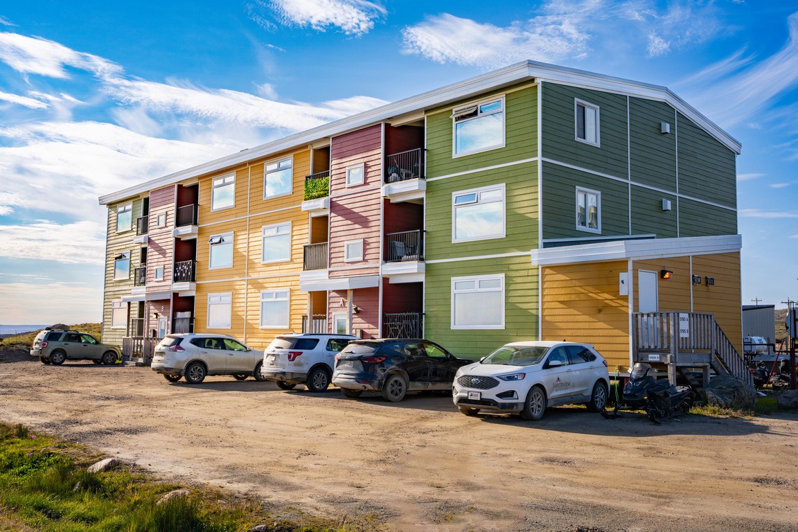 Colorful three-story apartment building with parked SUVs on a dirt lot under blue sky.