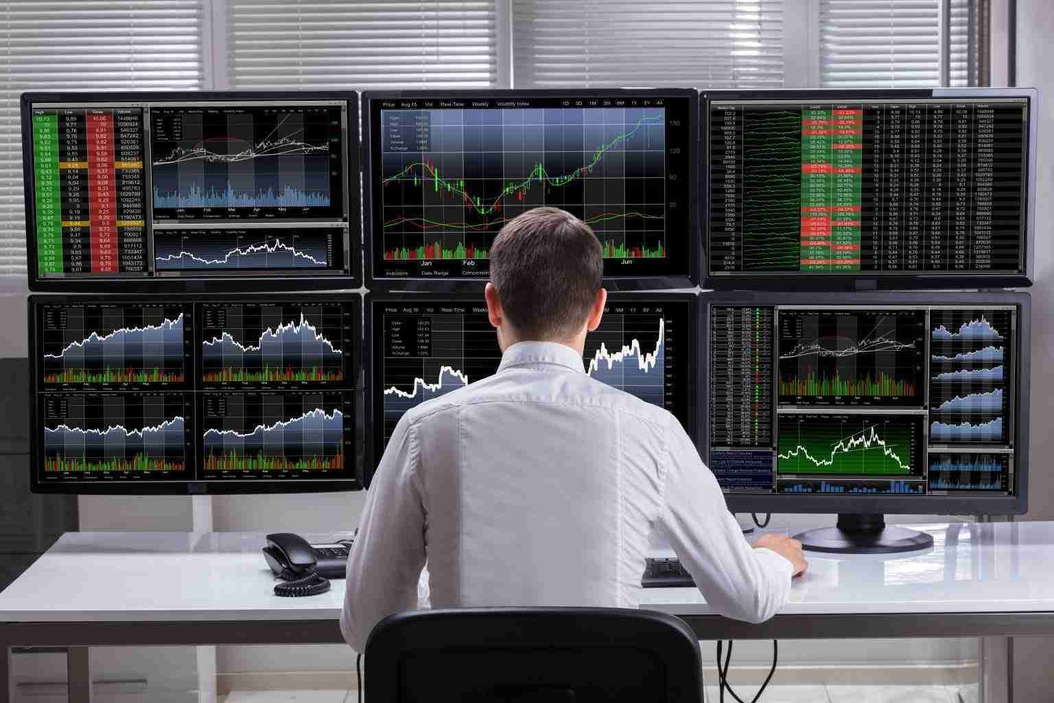 A man analyzes financial charts on multiple monitors in an office setting. A man analyzes financial charts on multiple monitors in an office setting.
