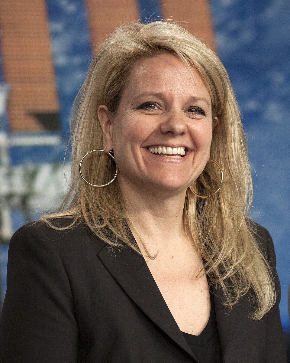 A blonde woman in a black blazer smiles against a blue-brown patterned backdrop. A blonde woman in a black blazer smiles against a blue-brown patterned backdrop.