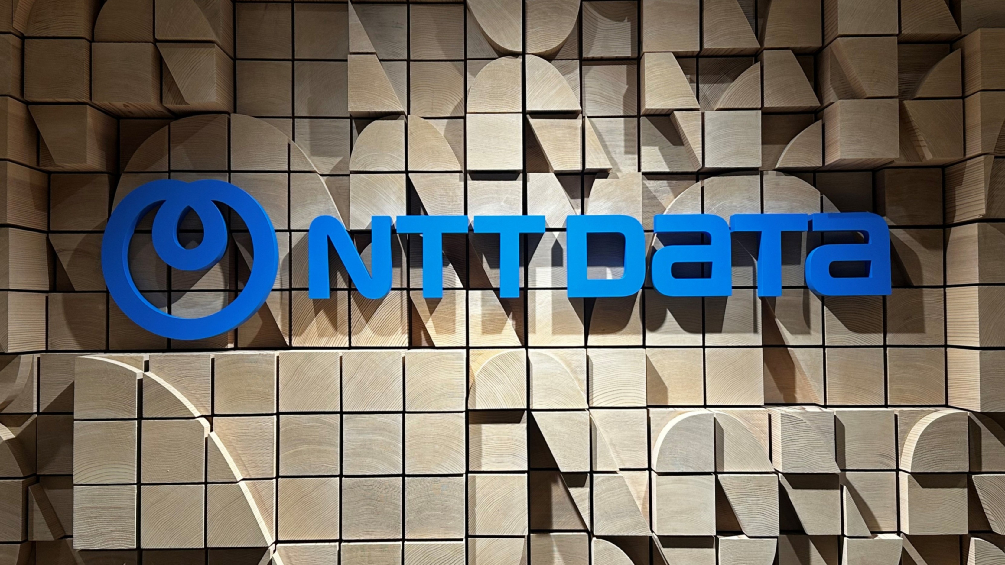 NTT DATA logo displayed on a geometric wooden panel wall. NTT DATA logo displayed on a geometric wooden panel wall.