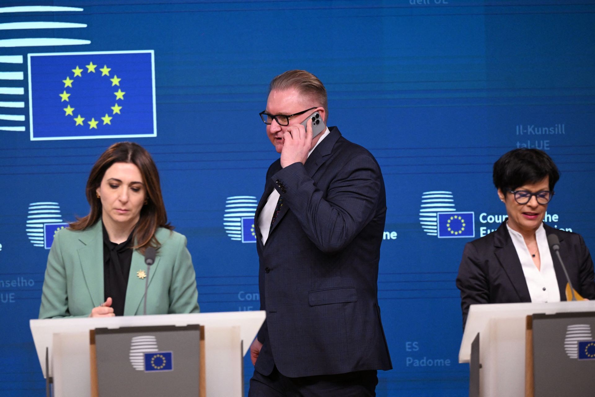 Man talks on mobile phone at EU press conference with two women at podiums. Man talks on mobile phone at EU press conference with two women at podiums.