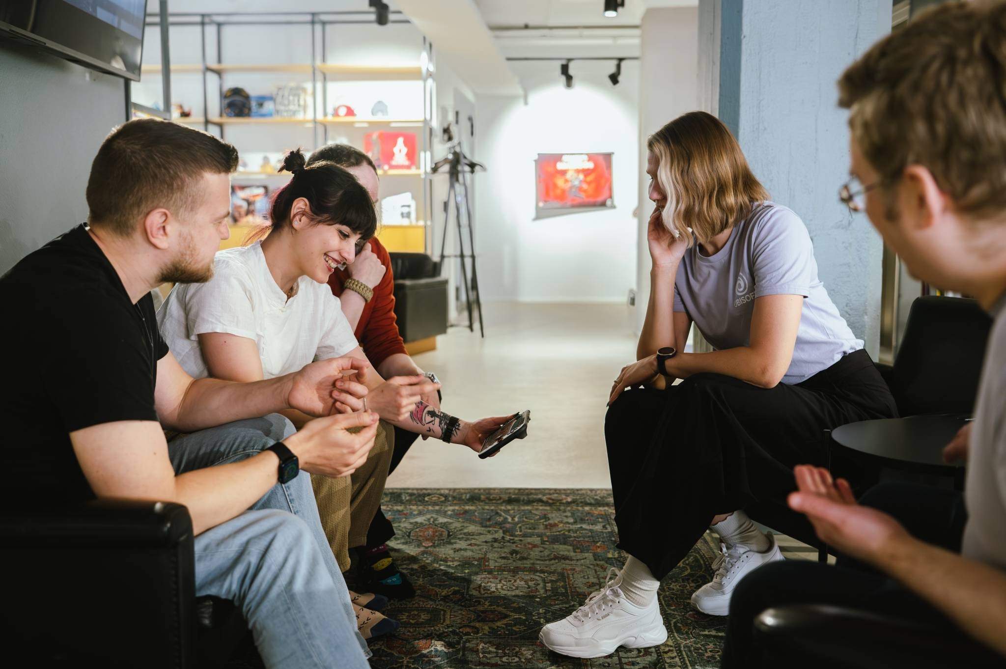 Five people sit in an open-plan office; one holds a phone while others interact. Five people sit in an open-plan office; one holds a phone while others interact.