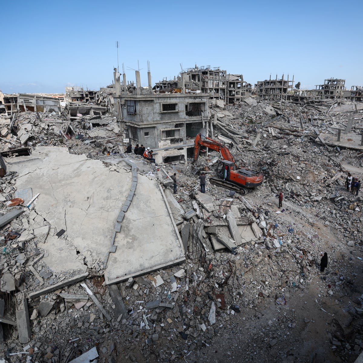 Workers and an excavator clear rubble from destroyed buildings in a devastated urban area. Workers and an excavator clear rubble from destroyed buildings in a devastated urban area.