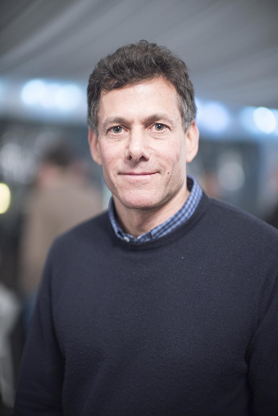 A man in a dark sweater stands facing camera indoors with blurred background lights. A man in a dark sweater stands facing camera indoors with blurred background lights.