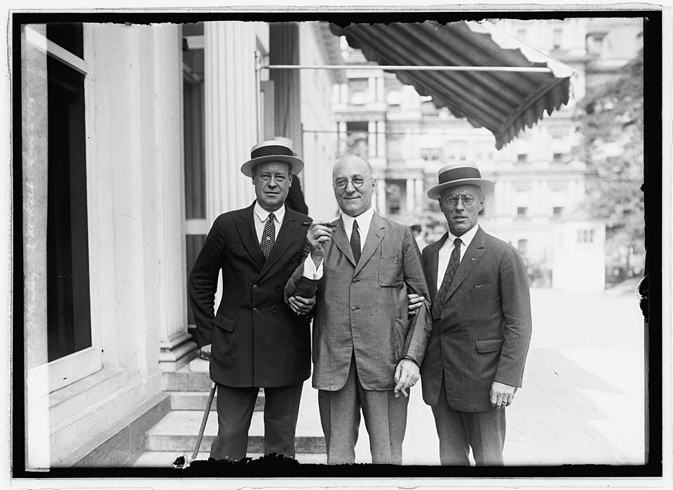 Three men pose for a photograph outside a columned building under an awning.