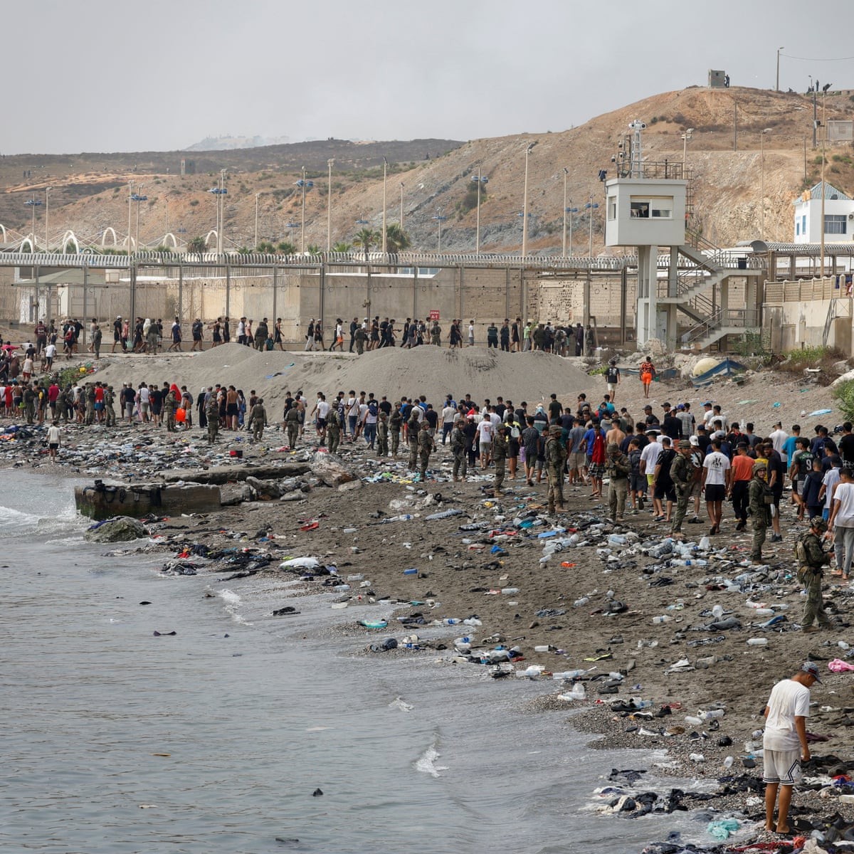 People gather on a debris-littered beach adjacent to a fenced coastal structure. People gather on a debris-littered beach adjacent to a fenced coastal structure.