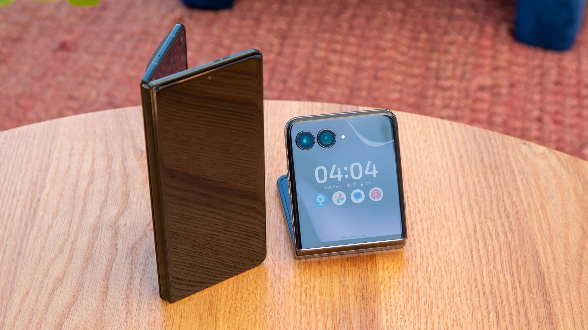 Foldable smartphone shows 04:04 on outer screen while one panel stands upright on a wooden table. Foldable smartphone shows 04:04 on outer screen while one panel stands upright on a wooden table.