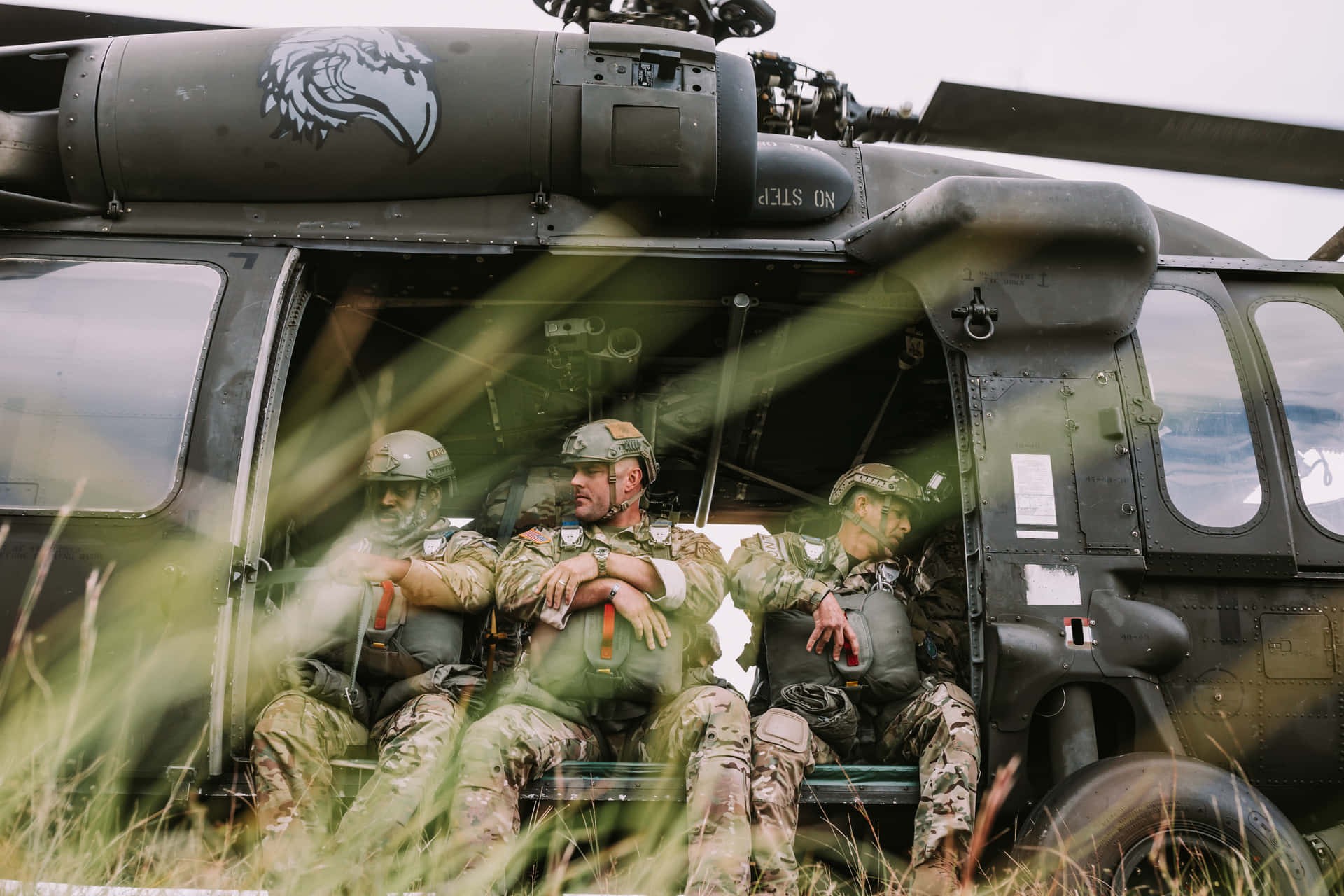 Three soldiers in camouflage uniforms sit in the open doorway of a military helicopter on grassy terrain. Three soldiers in camouflage uniforms sit in the open doorway of a military helicopter on grassy terrain.