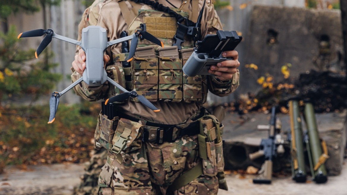 Soldier in camouflage uniform holds drone and controller outdoors near military equipment. Soldier in camouflage uniform holds drone and controller outdoors near military equipment.