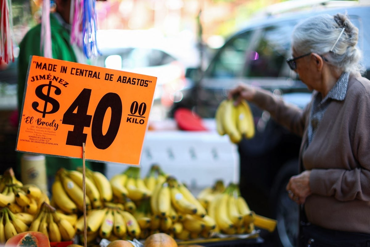 An elderly woman selects bananas at a street market with an orange "$40/kilo" price sign visible. An elderly woman selects bananas at a street market with an orange "$40/kilo" price sign visible.