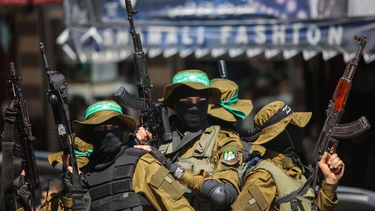 Armed men in olive uniforms and green headbands hold rifles outside an "IRALI FASHION" storefront. Armed men in olive uniforms and green headbands hold rifles outside an "IRALI FASHION" storefront.
