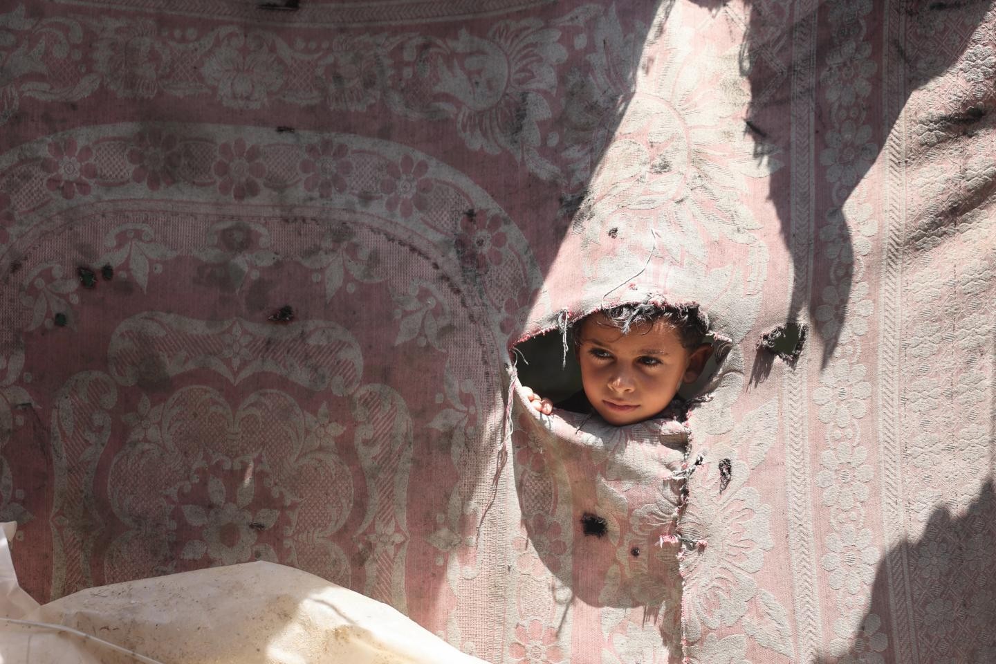 A child peers through a torn hole in a pink floral-patterned fabric wall.