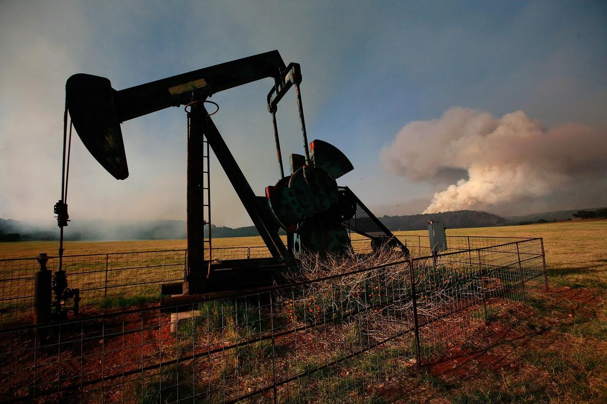 Oil pumpjack pumps in fenced field with smoke rising over distant hills.
