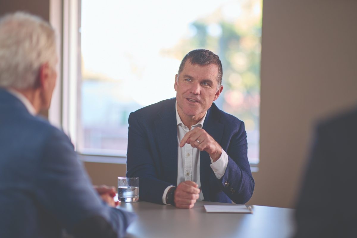 Man in blue suit speaks and gestures during a meeting at a table near a window. Man in blue suit speaks and gestures during a meeting at a table near a window.