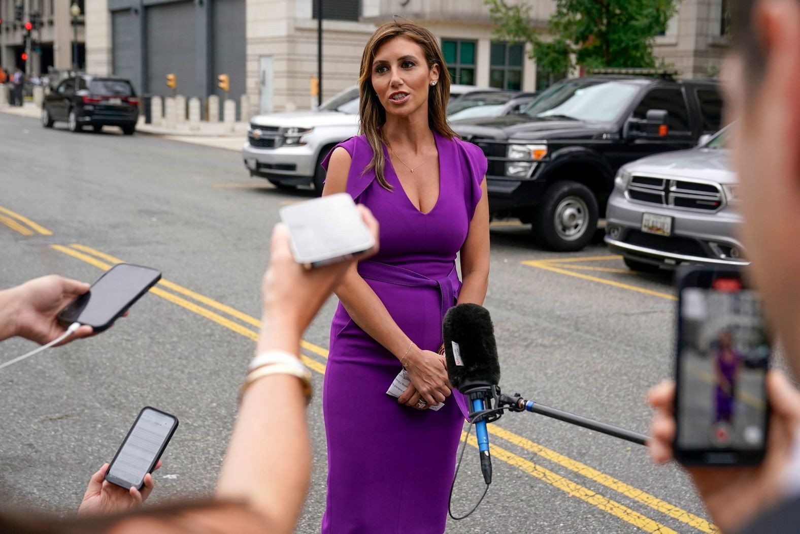 A woman in a purple dress is interviewed on a city street by journalists holding microphones and smartphones. A woman in a purple dress is interviewed on a city street by journalists holding microphones and smartphones.
