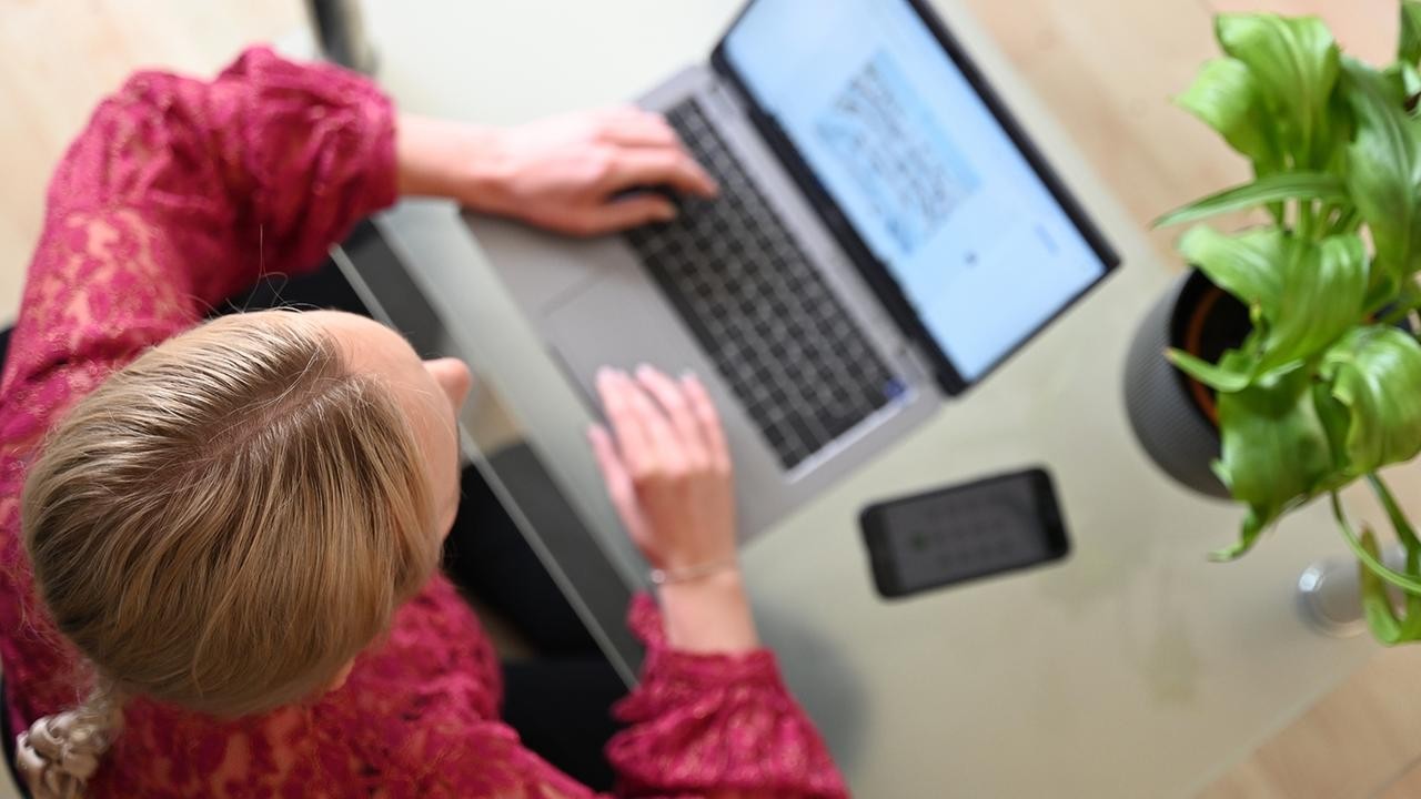 A woman in a red top types on a laptop at a glass table with a plant nearby. A woman in a red top types on a laptop at a glass table with a plant nearby.
