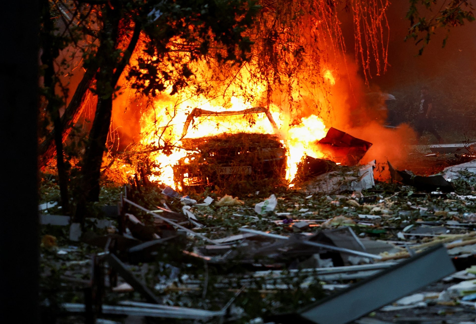 A vehicle burns in flames amid rubble and trees. A vehicle burns in flames amid rubble and trees.