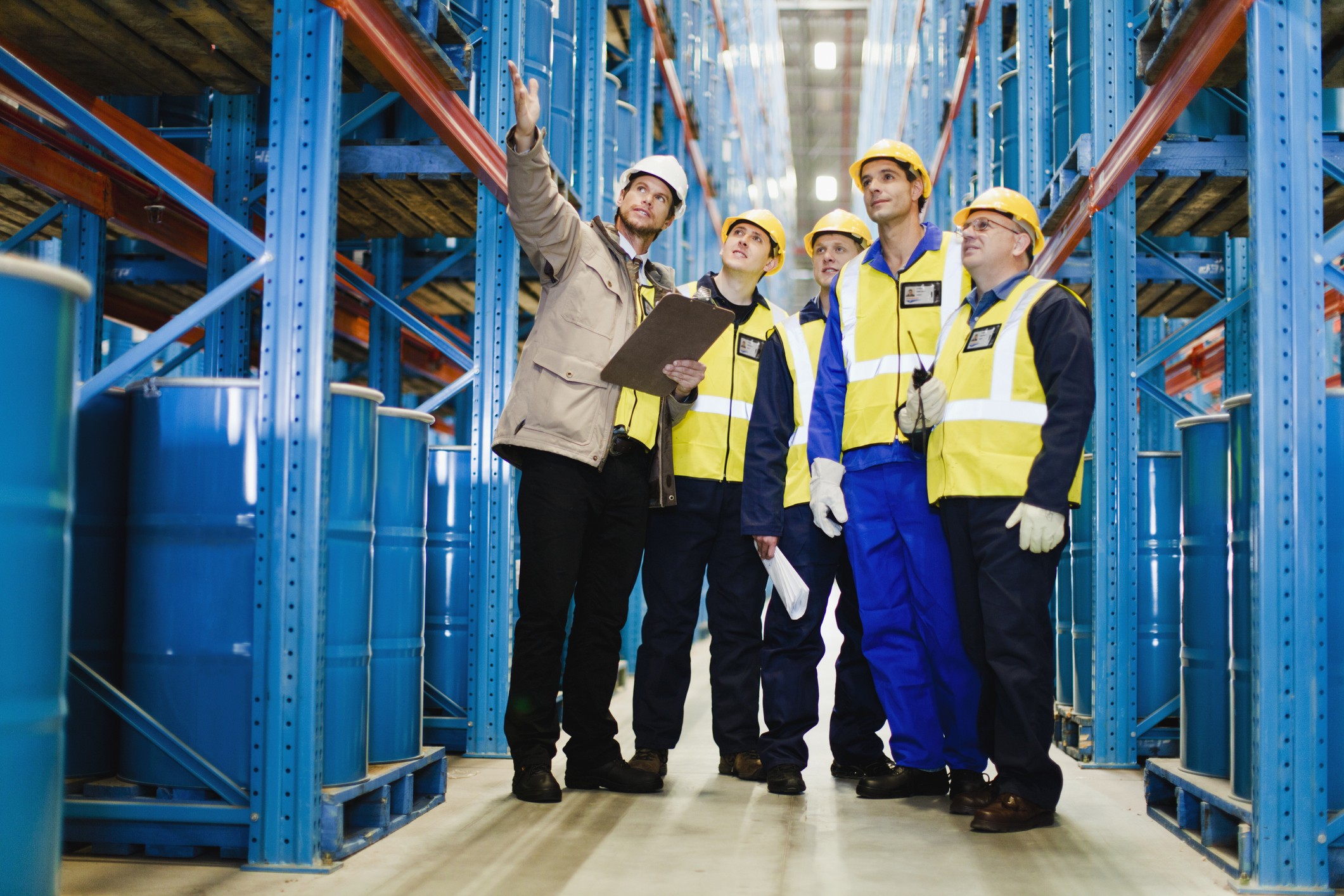 Workers in safety vests inspect warehouse storage racks; one gestures upward while others observe. Workers in safety vests inspect warehouse storage racks; one gestures upward while others observe.