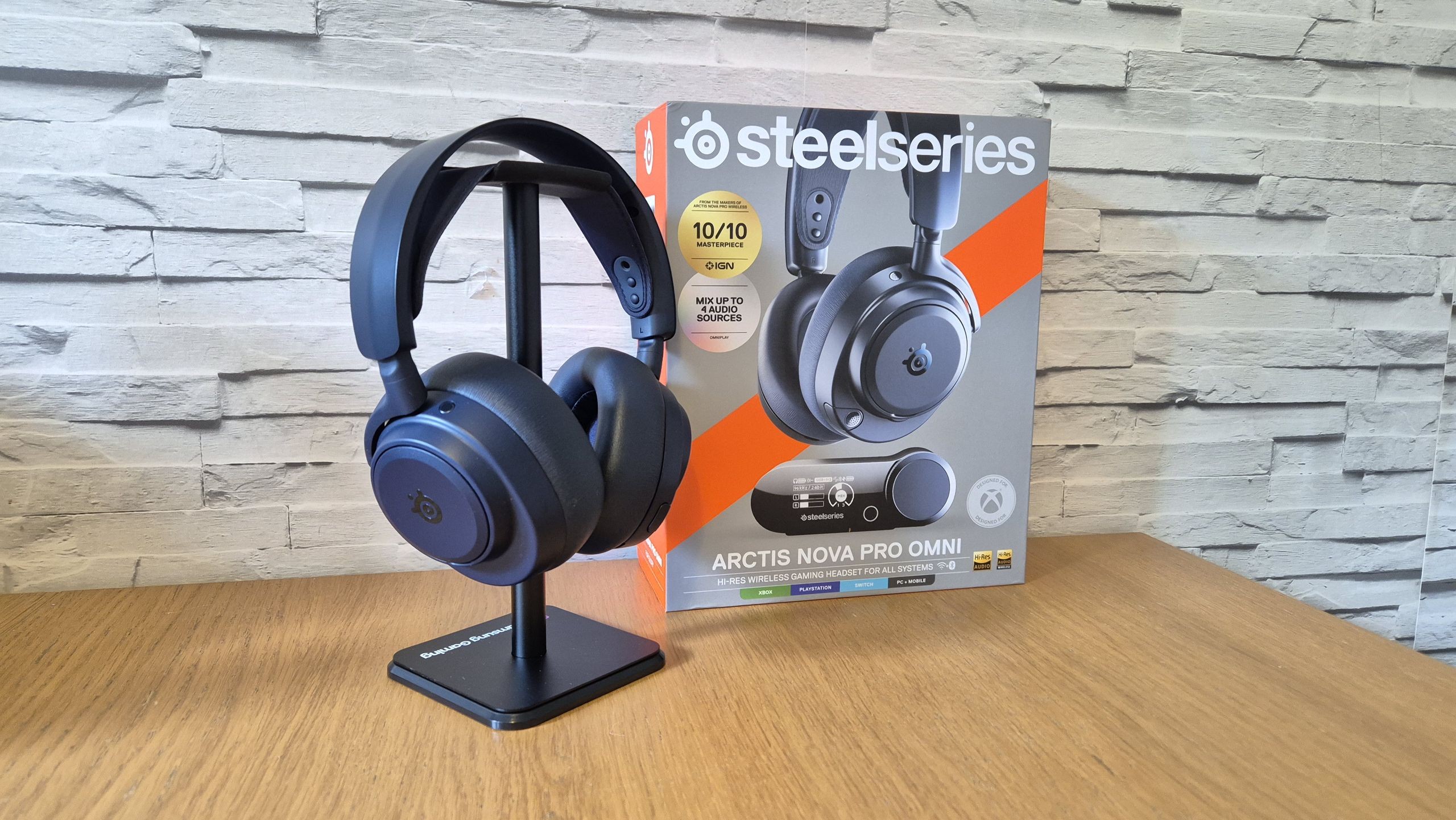SteelSeries Arctis Nova Pro Omni headset on display stand beside its box on wooden table against brick wall. SteelSeries Arctis Nova Pro Omni headset on display stand beside its box on wooden table against brick wall.