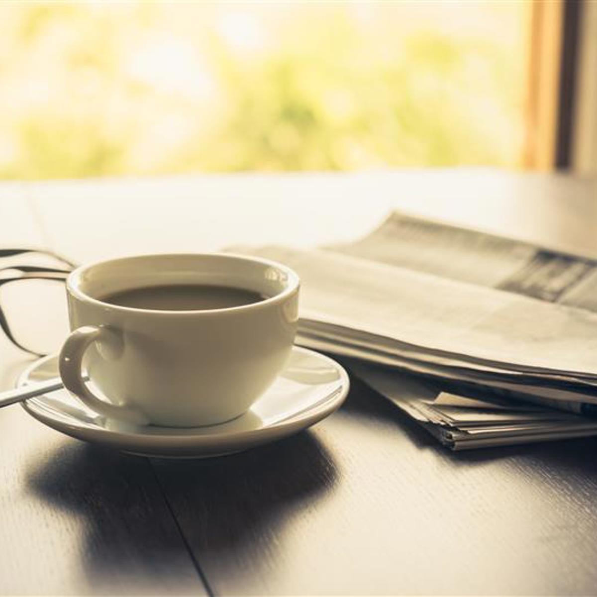 A white coffee cup on a saucer sits beside newspapers and eyeglasses on a wooden table near a window. A white coffee cup on a saucer sits beside newspapers and eyeglasses on a wooden table near a window.
