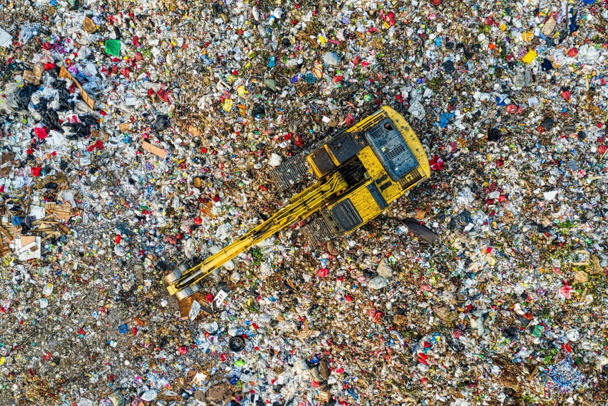 A yellow excavator operates within a vast landfill filled with mixed waste materials. A yellow excavator operates within a vast landfill filled with mixed waste materials.
