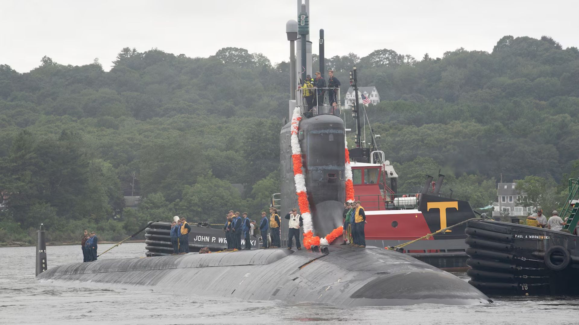 A submarine is towed through a river by tugboats with crew members visible. A submarine is towed through a river by tugboats with crew members visible.