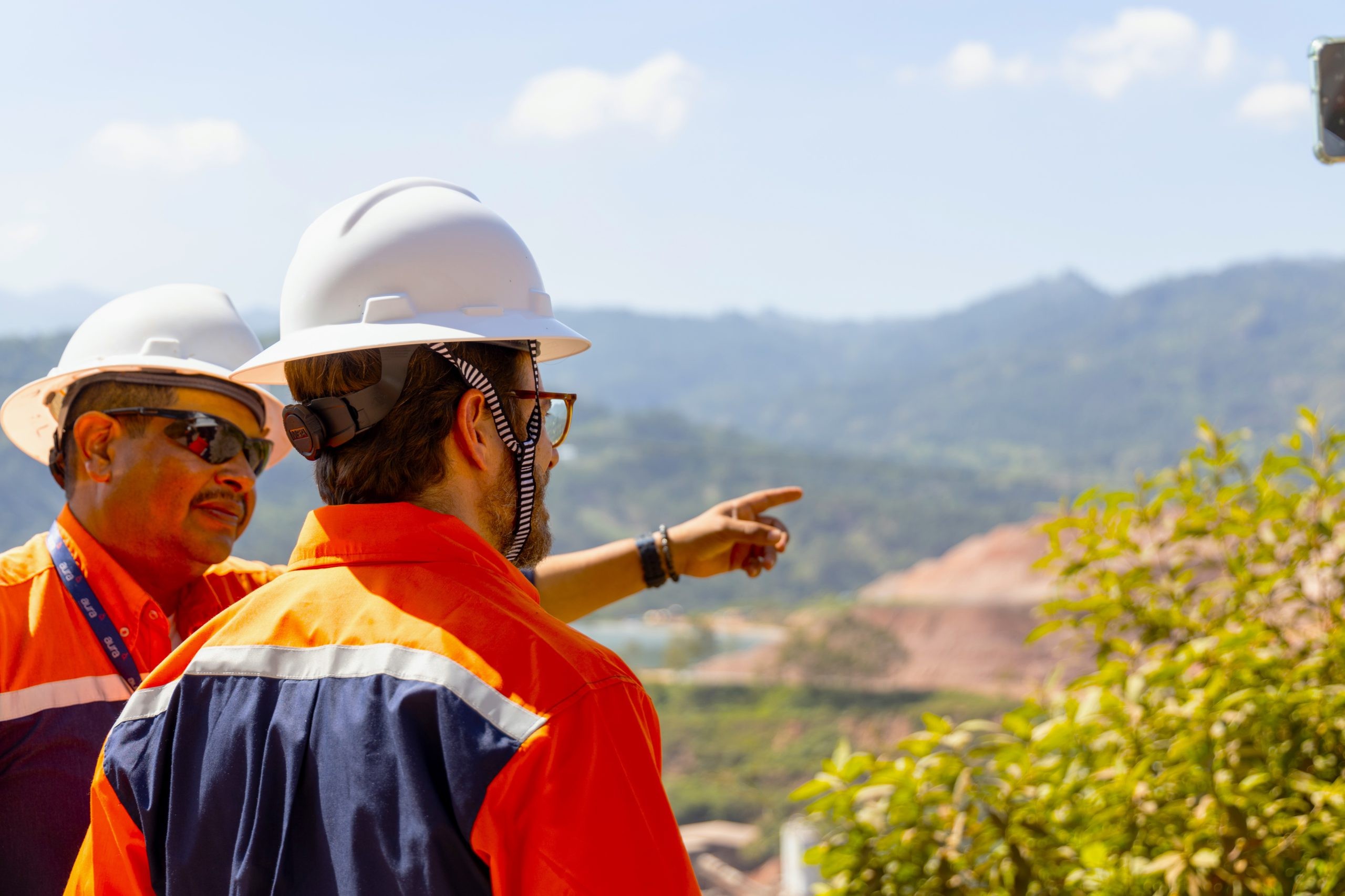 Two construction workers in hard hats point toward distant mountains outdoors.