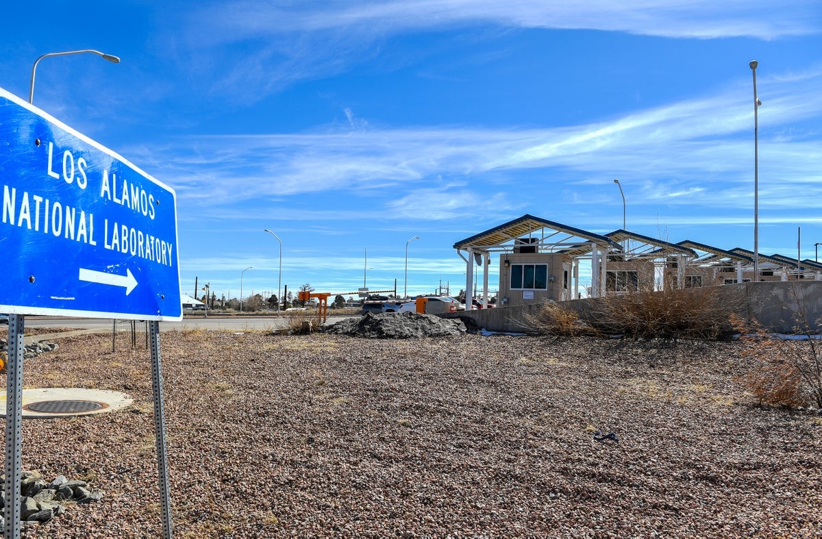 Blue "Los Alamos National Laboratory" sign points right on gravel lot near buildings. Blue "Los Alamos National Laboratory" sign points right on gravel lot near buildings.
