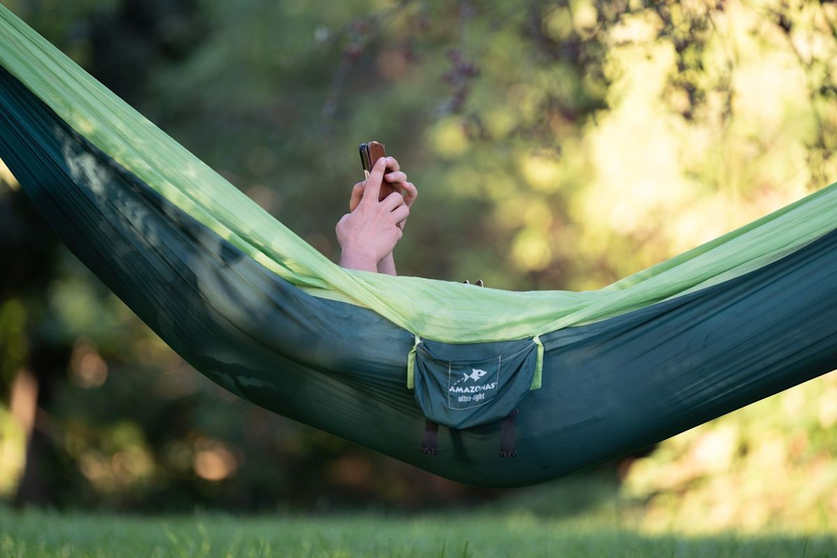 Person holds smartphone while reclining in a green hammock amid outdoor foliage. Person holds smartphone while reclining in a green hammock amid outdoor foliage.
