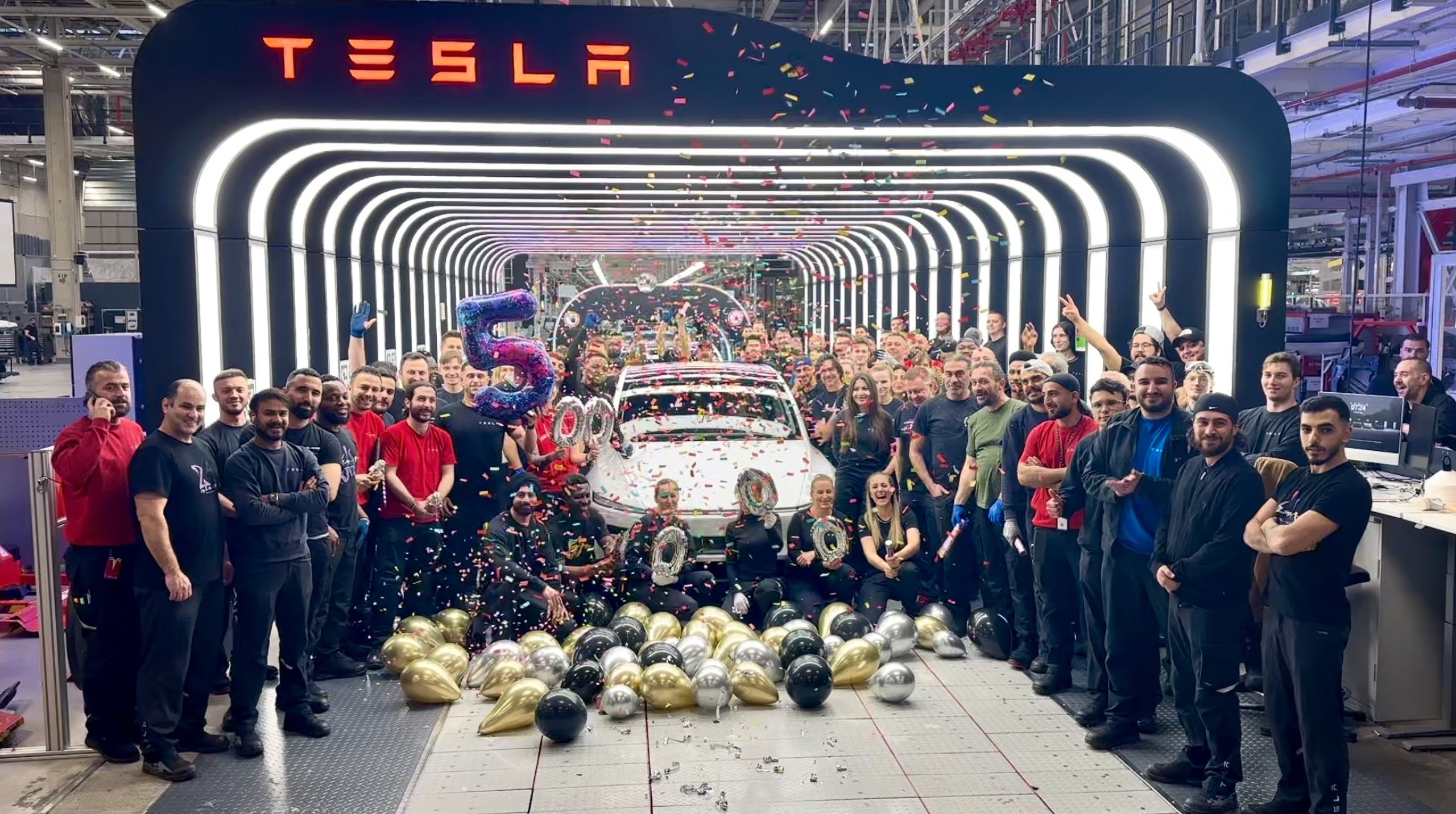 Tesla employees celebrate a production milestone in a factory tunnel with confetti and balloons. Tesla employees celebrate a production milestone in a factory tunnel with confetti and balloons.