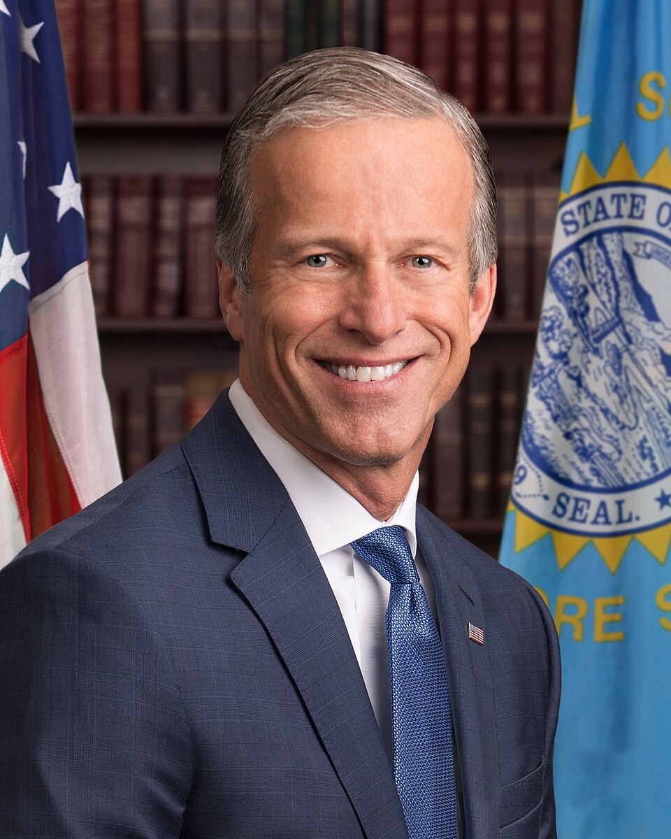 Man in blue suit smiles before American and state flags with bookshelves behind him. Man in blue suit smiles before American and state flags with bookshelves behind him.