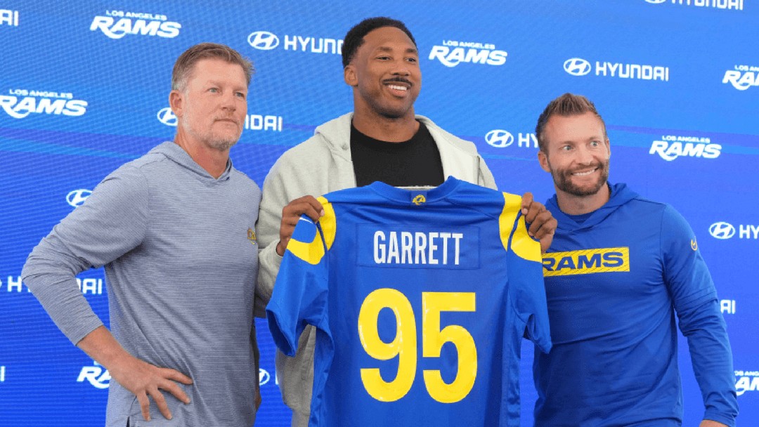 Three men hold a blue Los Angeles Rams jersey with "GARRETT" and #95 at a branded press event. Three men hold a blue Los Angeles Rams jersey with "GARRETT" and #95 at a branded press event.