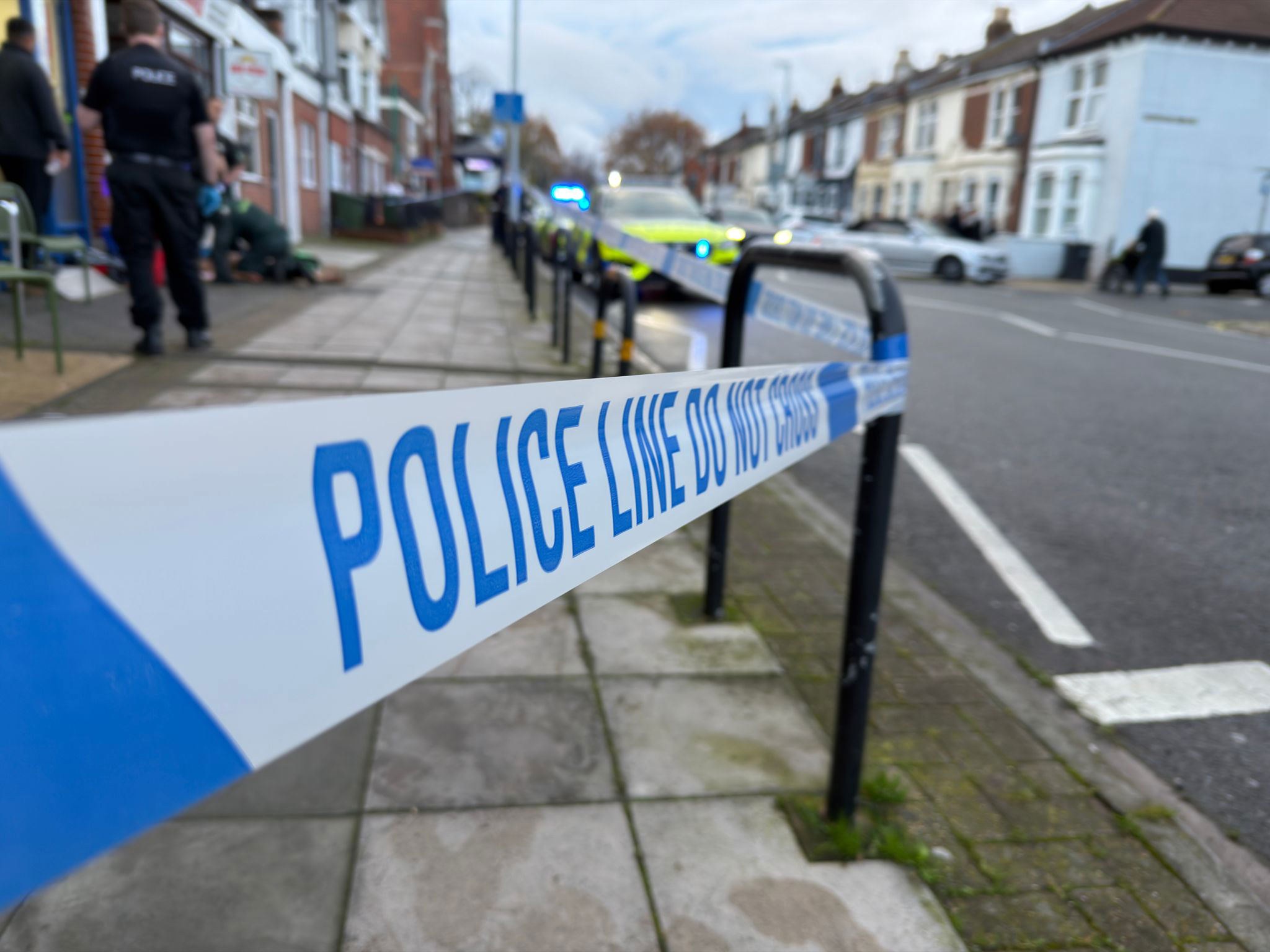 Police tape marks a police line on a residential sidewalk with officers and vehicles visible. Police tape marks a police line on a residential sidewalk with officers and vehicles visible.