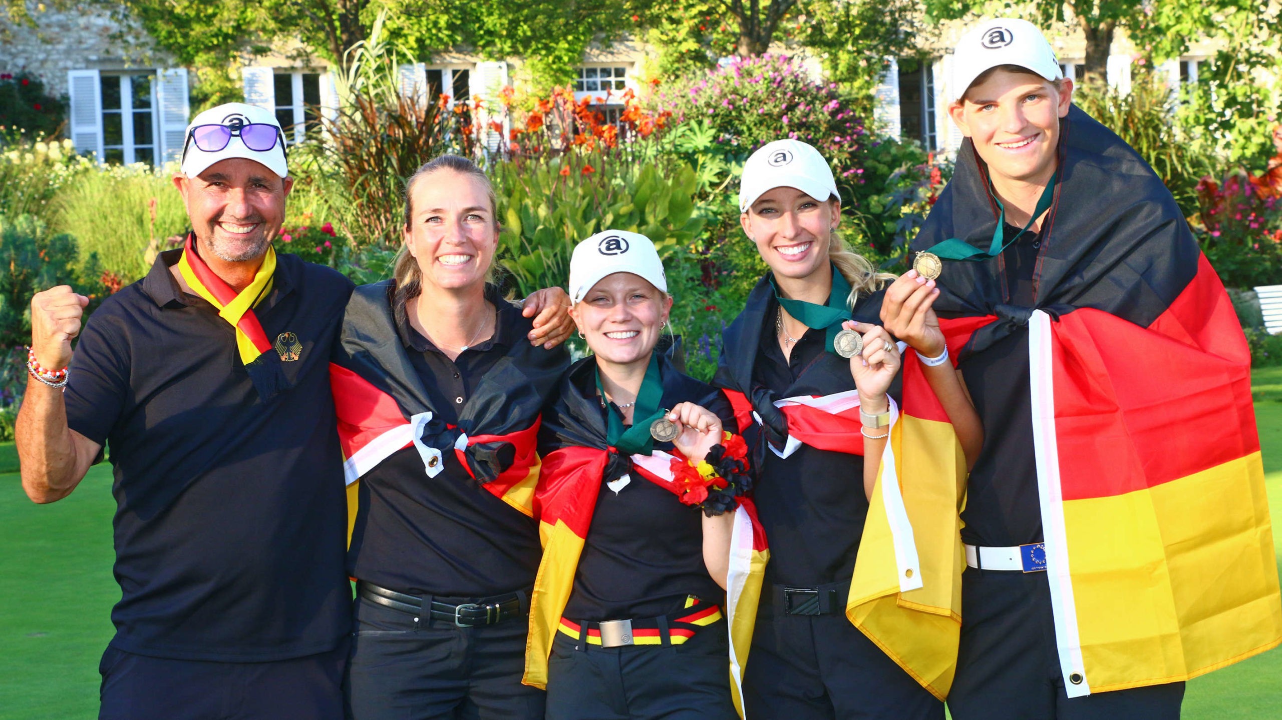 Five people draped in German flags hold medals and pose together in a garden setting.