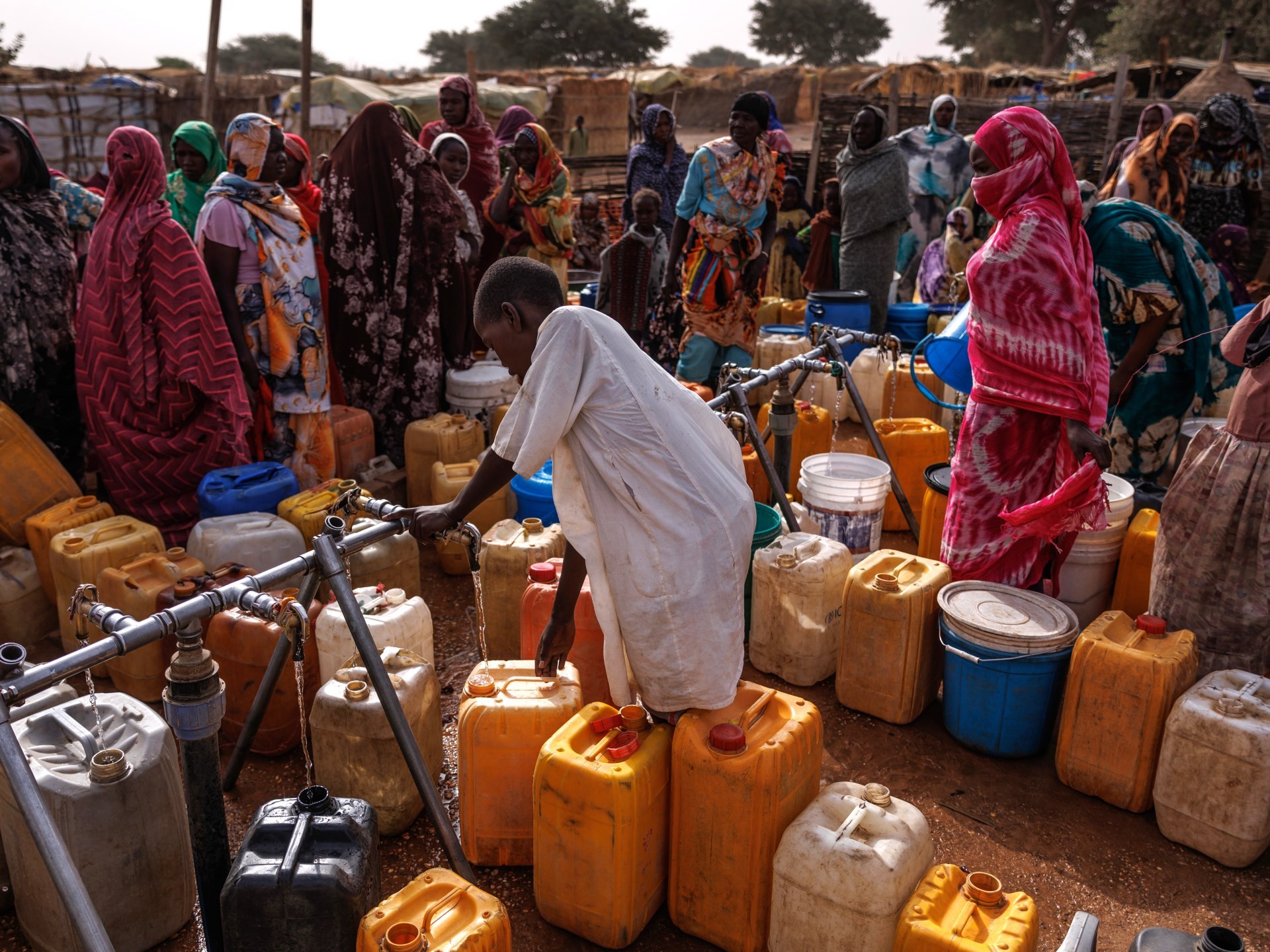 Boy fills yellow and white jerry cans at a public water point with onlookers. Boy fills yellow and white jerry cans at a public water point with onlookers.