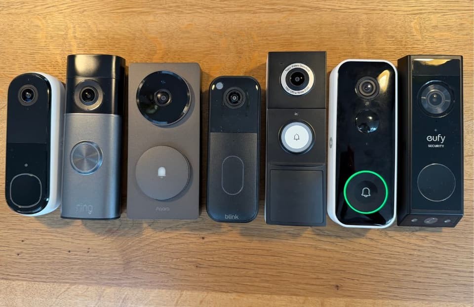 Two video doorbells mounted: white on textured wall with plant, black on wooden surface. Two video doorbells mounted: white on textured wall with plant, black on wooden surface.
