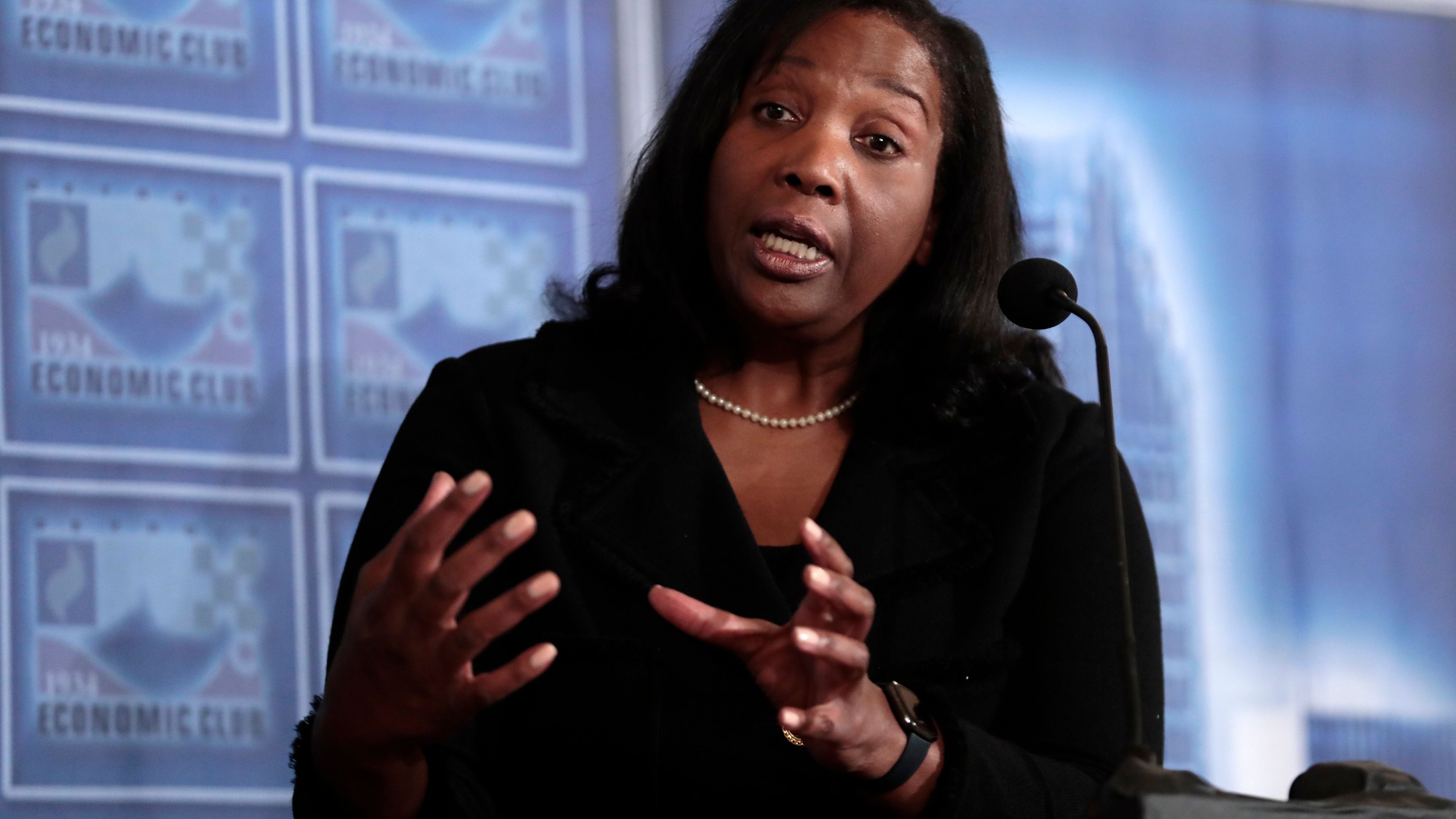Woman speaks into a microphone at an Economic Club event, gesturing with her hands.