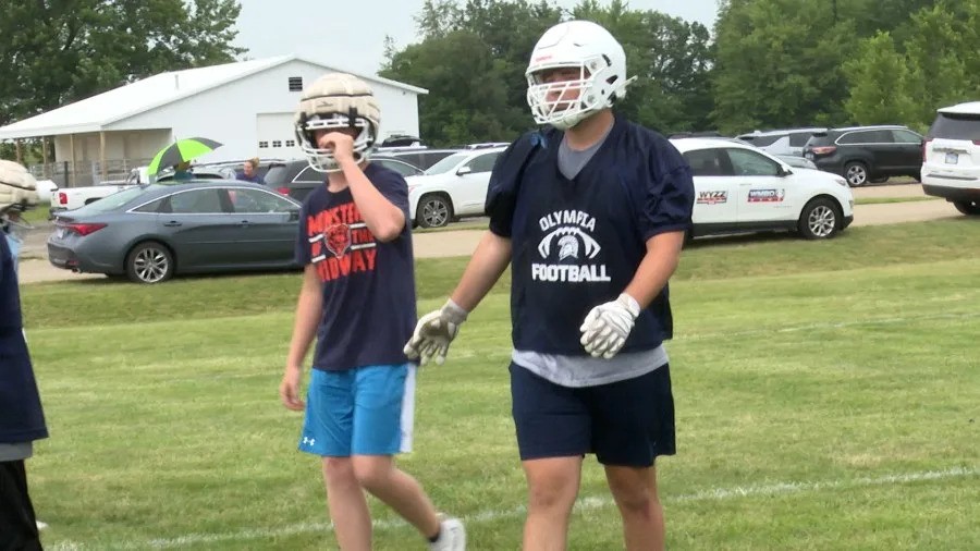 Two youth football players walk on a grassy field with parked cars and buildings in background. Two youth football players walk on a grassy field with parked cars and buildings in background.